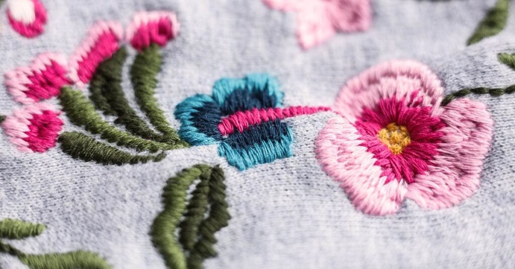 Bordado con lana sobre tela gris con flores en tonos rosados, azules y verdes, destacando la textura del hilo.